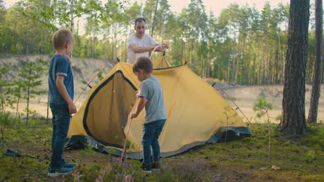 The-young-man-and-two-young-boys-together-set-up-a-tent-camp-for-the-night-during-the-campaign.-Fatherhood-and-a-happy-childhood.-Father-and-sons-put-up-a-tent-together-in-the-woods